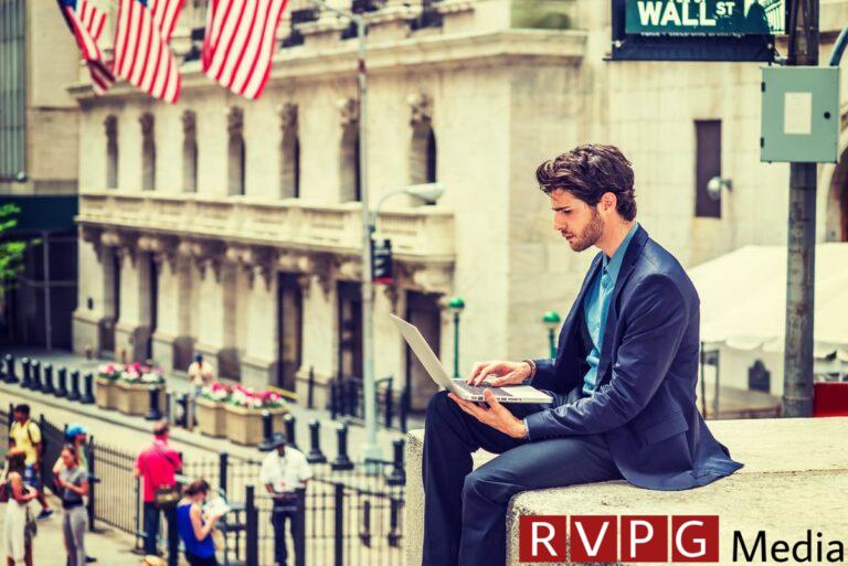 A man sitting on a ledge looking at his laptop. The street sign above him reads Wall St.